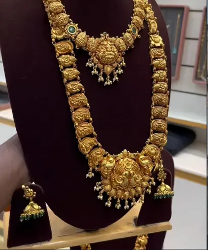 An antique temple haram with carved medallions and green stone drops, with matching jhumkas, on a maroon bust.
