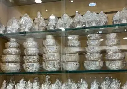 Glass shelves stacked with silver vessels, bowls and pooja articles.